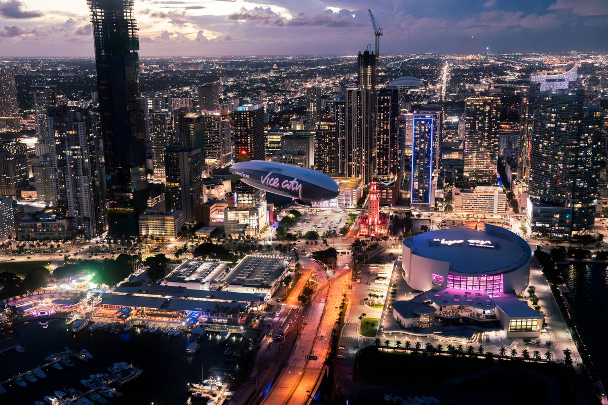 Vice City branded Goodyear blimp above Miami at dusk, with the waterfront arena and city lights below.